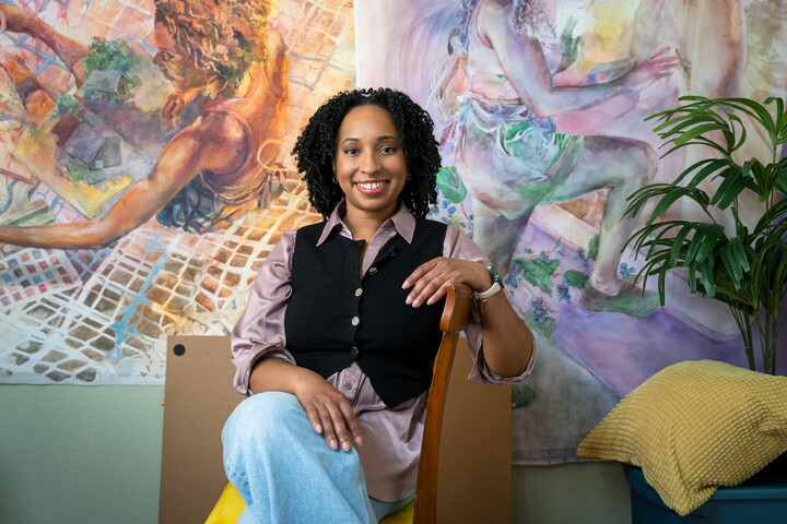A smiling Black woman with curly hair is seated in a chair in front of several large-scale paintings.