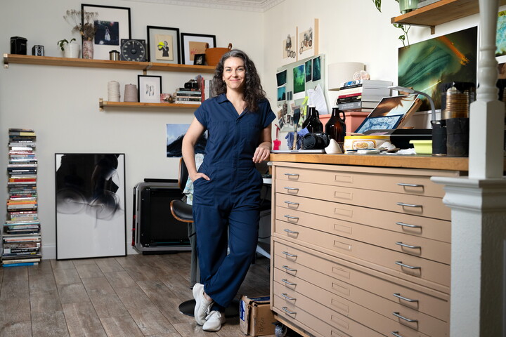 A White woman with dark curly hair wearing a blue jumpsuit leans against an art file cabinet in a studio.