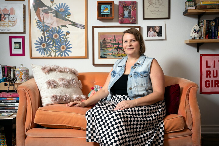 A White woman with short blonde hair is seated on an orange loveseat surrounded by books and artwork.