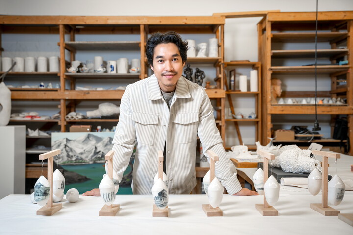 A medium-skinned man with Asian features leans against a table in a workshop, on which is displayed groupings of ceramic sculptures.