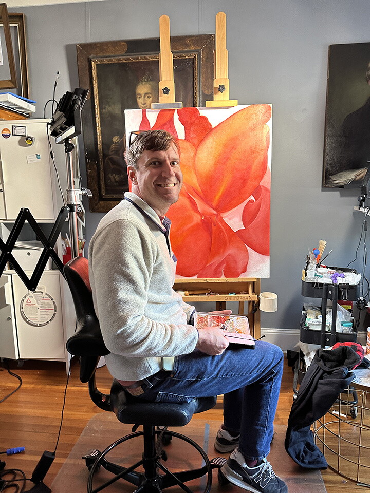 A White man sits in front of a painting of red flower petals that is resting on an easel.