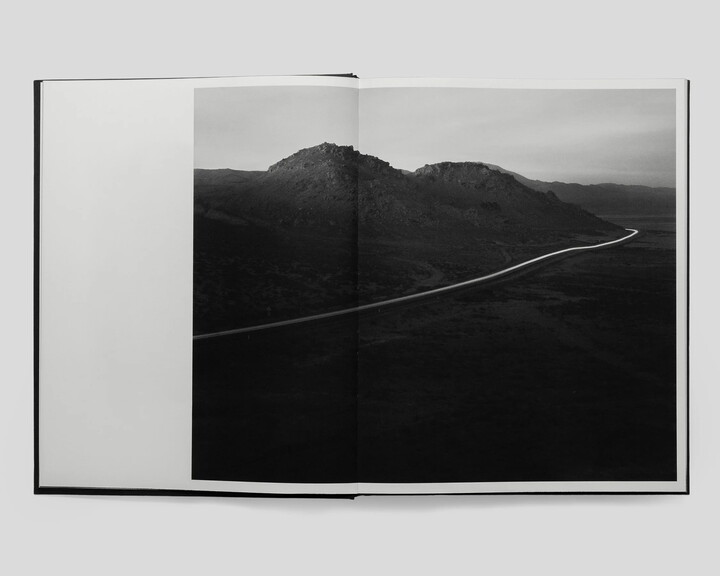 An open book with a black-and-white photograph spanning both pages of a lonely road bisecting the side of dark hills.