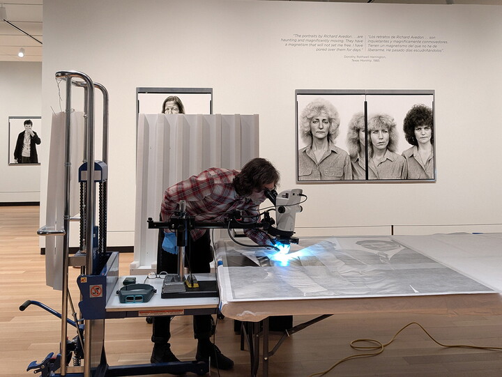 A White man leans over a microscope-like piece of equipment to inspect a large-scale photograph that is laying on a table in a Carter gallery.