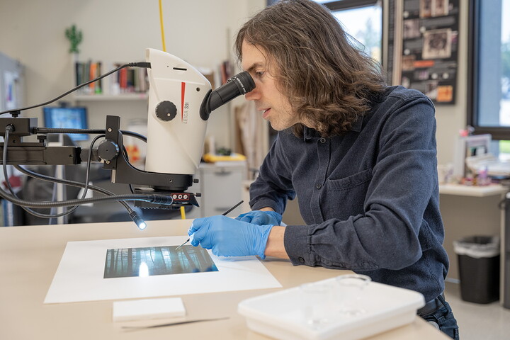 A White man with longish dark hair looks into a microscope-like device to view details on the photograph beneath it.
