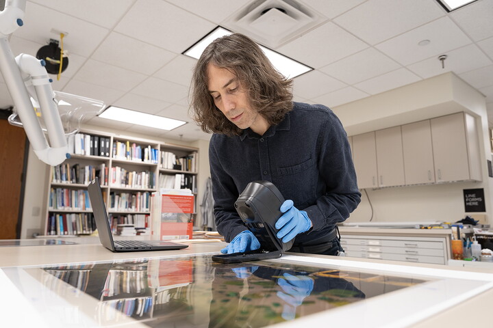 A White man with longish dark hair holds a piece of equipment, hands covered in latex gloves, over a large photograph laying on a table.