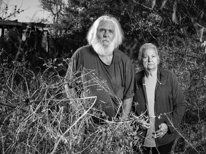 A black-and-white photograph of two people standing in tall grass: one the left, an older man with white hair and beard, on the right, an older woman.