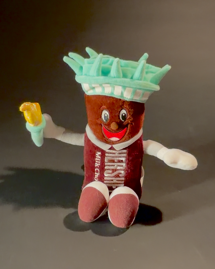 A plush Hershey bar with arms and legs wearing a Statue of Liberty crown and holding a torch.
