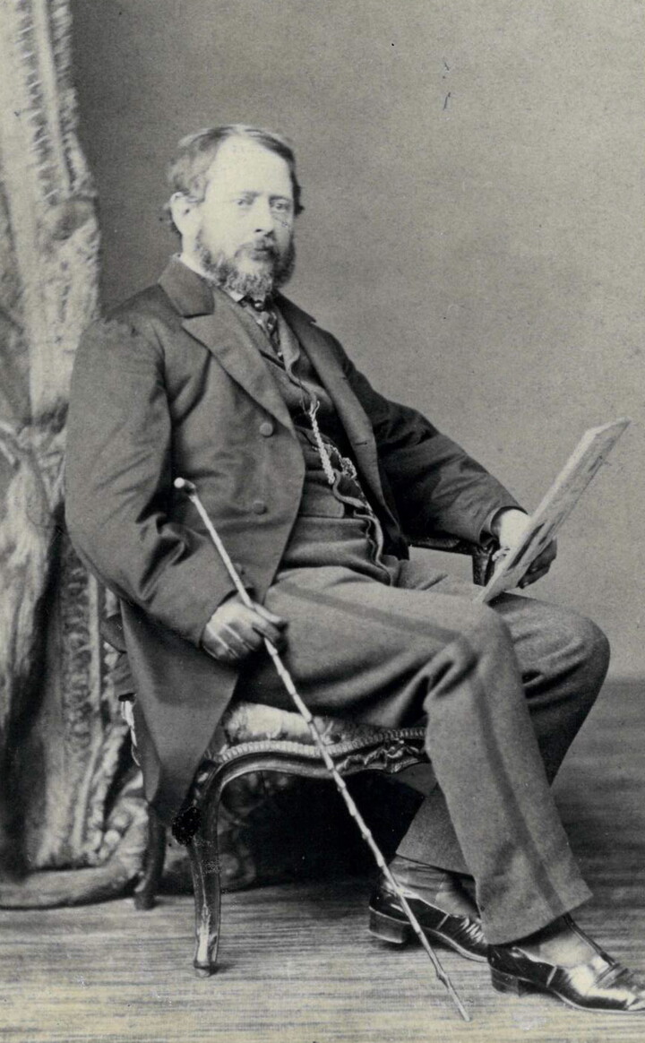 A black-and-white portrait photograph of a seated middle-aged White man with a beard and mustache wearing a three-piece suit, a cane resting by his side.