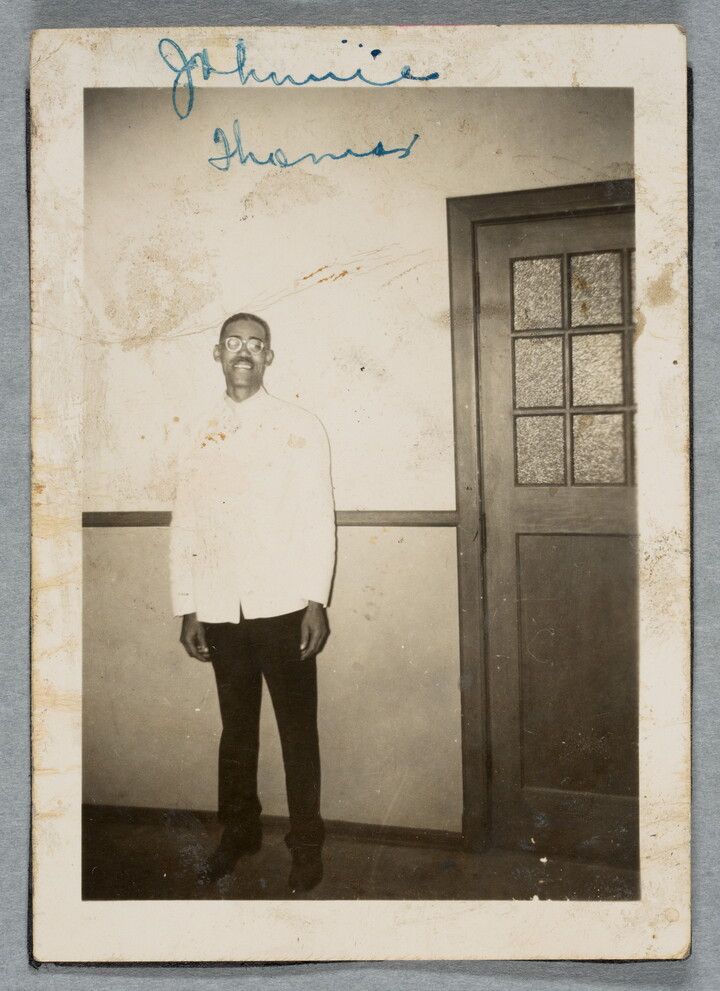 A black-and-white photograph of a Black man with short hair, glasses, a mustache, wearing a white jacket and black pants posing next to an interior door.