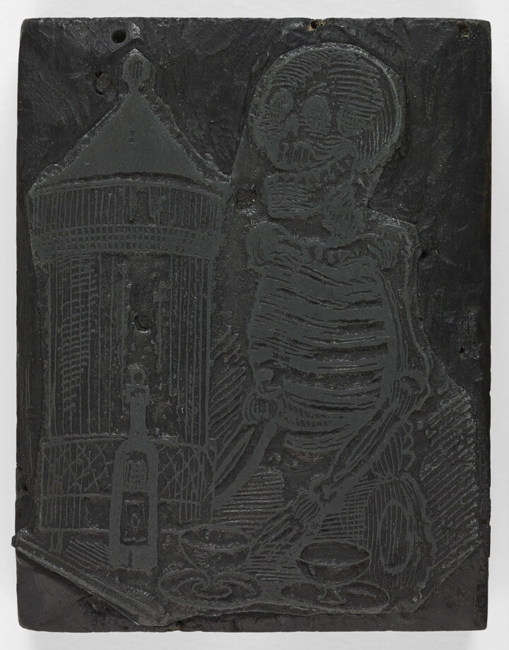 An engraved wood printing block depicting a skeleton standing in front of a table with a bottle and two glasses.
