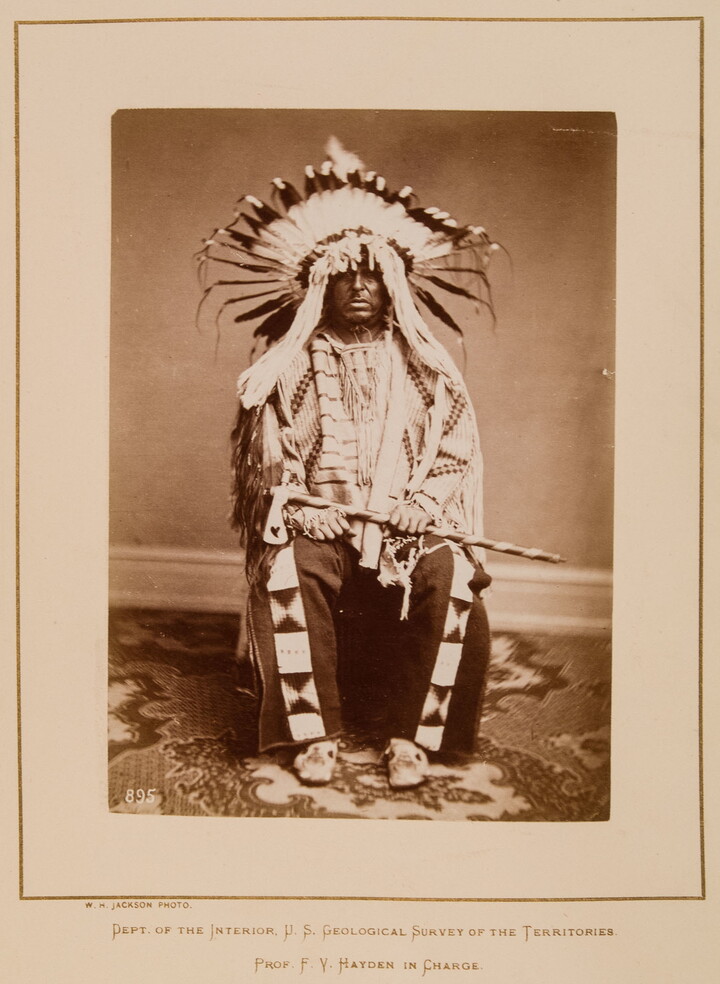 A black-and-white formal portrait photograph of a seated Indigenous man in full, traditional regalia with a headdress and tomahawk.