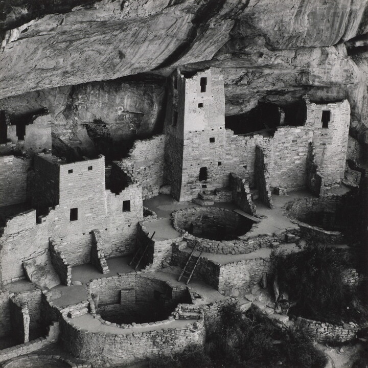 A black-and-white photograph of ruined stone buildings, some with towers, and several underground circular rooms all under the overhang of a cliff.