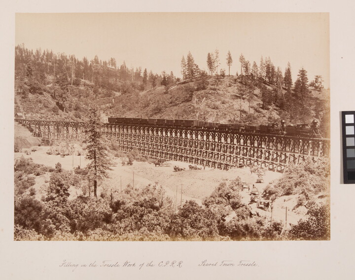 A sepia-toned photograph of a train travelling across a mountain valley with onlookers at base of the trestle.