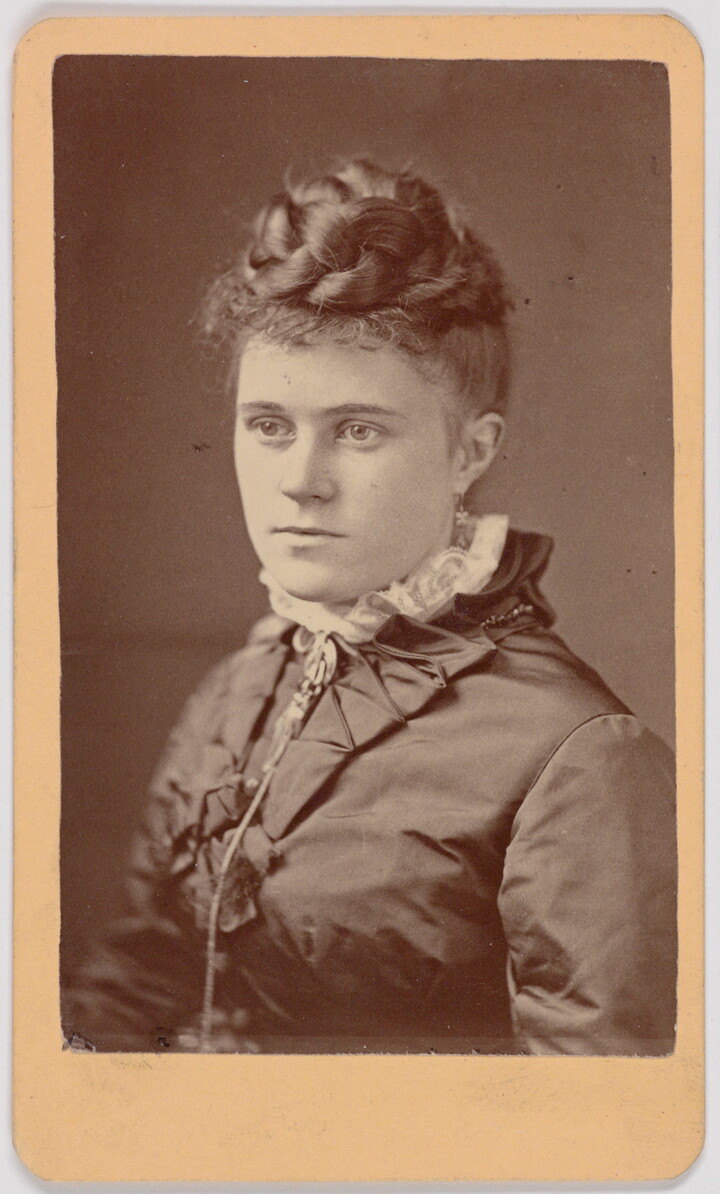 A black-and-white portrait photograph of a young White woman, hair braided and piled on top of her head, wearing a dark dress with a high lace collar.