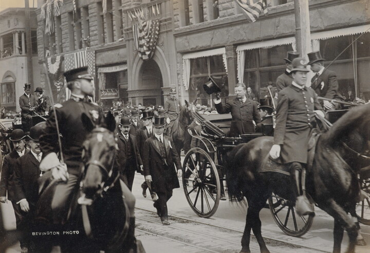 A black-and-white photograph of a parade of people on horseback, with a White man in the center in a carriage tipping his hat to onlookers.