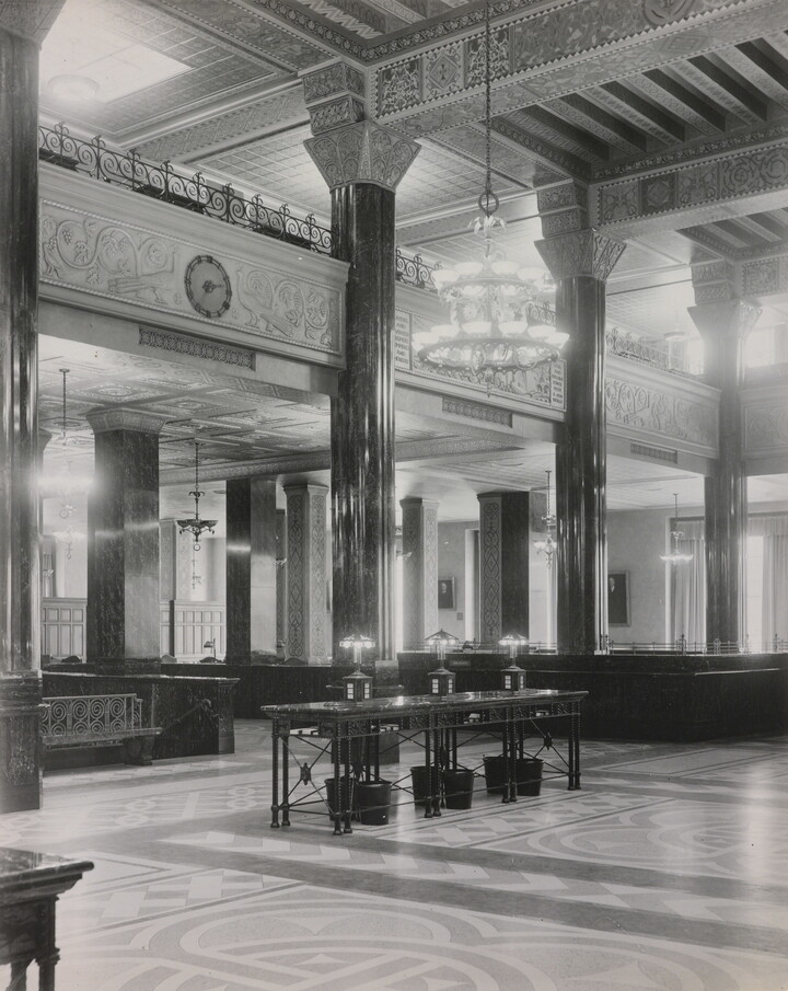 A black-and-white photograph of the interior of an ornate office lobby with inlaid floors, columns, and chandeliers.
