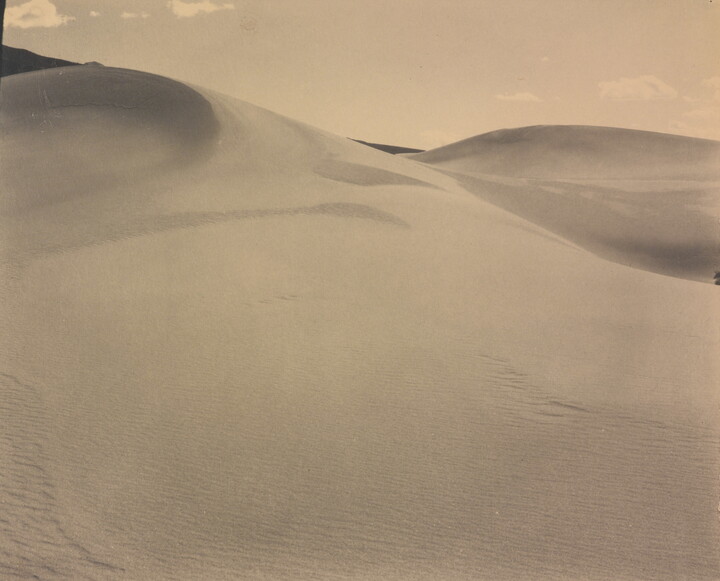 A sepia-toned photograph of a rippling sand dune rising up into a sky dotted with a few small clouds.