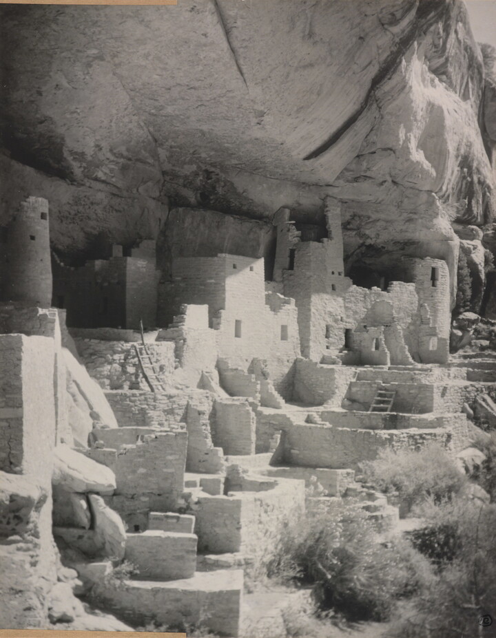 A black-and-white photograph of an abandoned, crumbling rock city built into the side of a desert cliff.