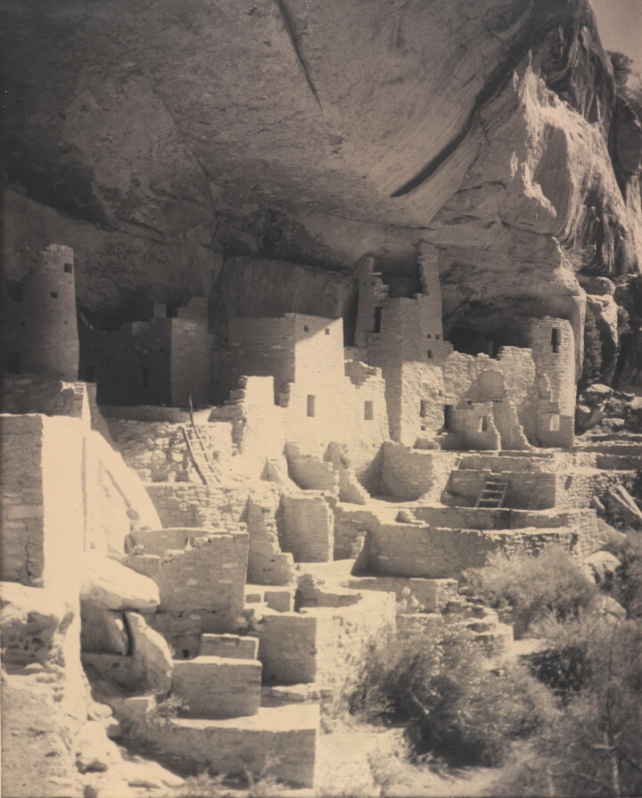 A black-and-white photograph of an abandoned, crumbling rock city built into the side of a desert cliff.