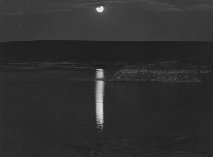 A black-and-white photograph of a night sky and full moon reflected across calm water.