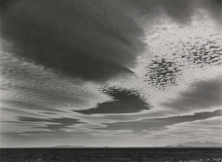 A black-and-white photograph of an expansive desert plain under rippled clouds with mountains in the far distance.
