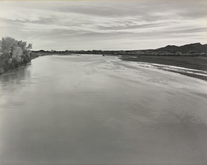 A black-and-white photograph of a calm river with trees on the left shore and mountains in the distance.