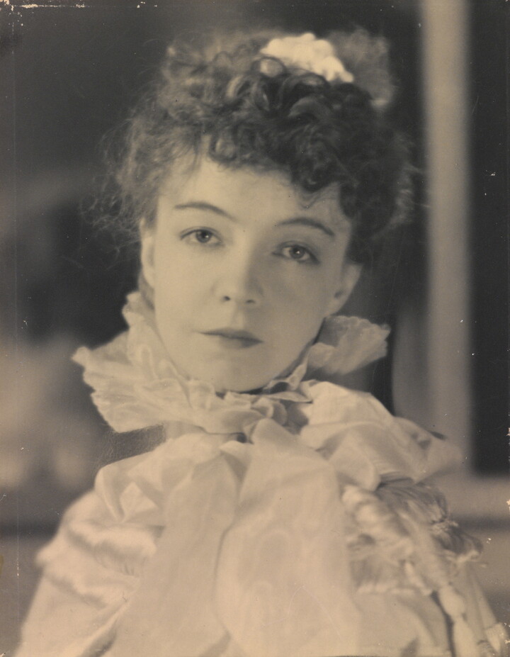 A black-and-white portrait photograph of a White woman with curly hair pulled on top of her head, wearing a top with a ruffled collar.