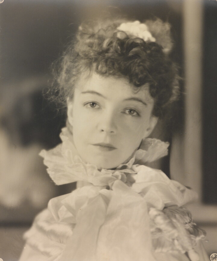 A black-and-white portrait photograph of a White woman with curly hair pulled on top of her head, wearing a top with a ruffled collar.