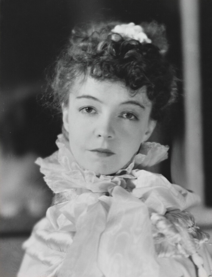 A black-and-white portrait photograph of a White woman with curly hair pulled on top of her head, wearing a top with a ruffled collar.