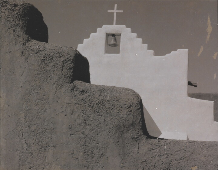 A black-and-white photograph of a terraced adobe bell tower with a cross on top, partially hidden by a terraced adobe wall.