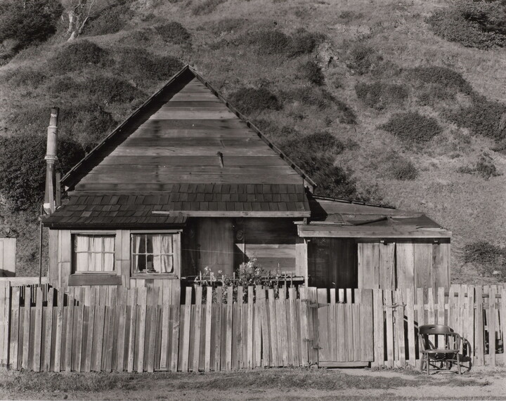 A black-and-white photograph of a small clapboard house behind a picket fence with a chair next to the gate.
