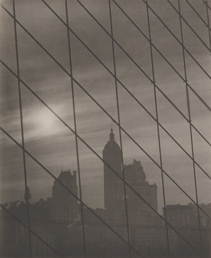 A black-and-white photograph of a grid of cables framing a cityscape with tall buildings in the background.