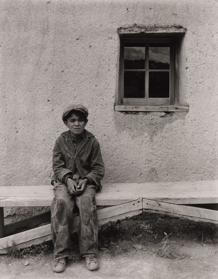 A black-and-white photograph of a squinting medium-skinned boy wearing a cap, jacket, and pants with patches on the knees, seated on a wooden bench in front of an adobe building.