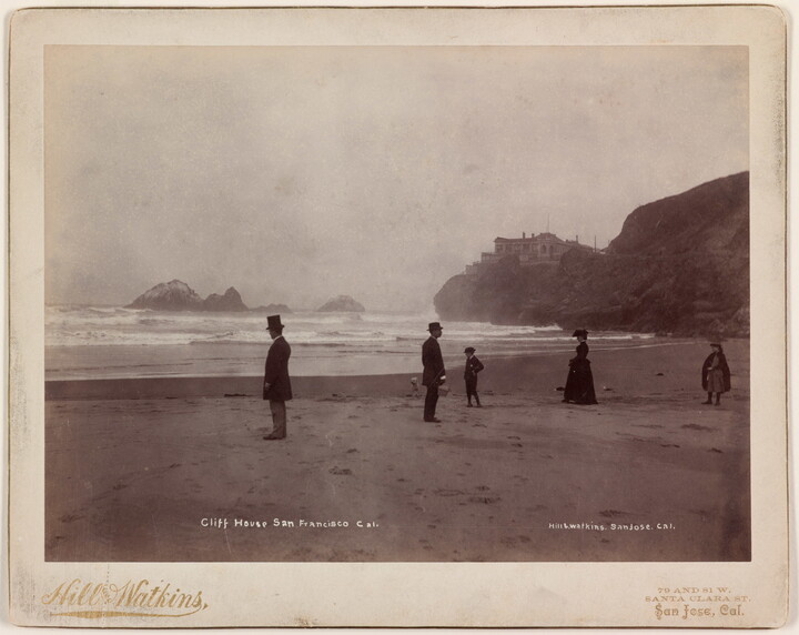 A black-and-white photographs of formally-dressed people scattered along a flat beach and calm shoreline; in the background, a building sits atop a high cliff.