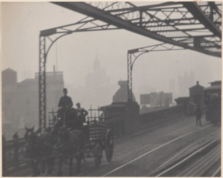 A black-and-white photograph of a horse-drawn carriage crossing a bridge next to streetcar rails as haze obscures buildings in the background.