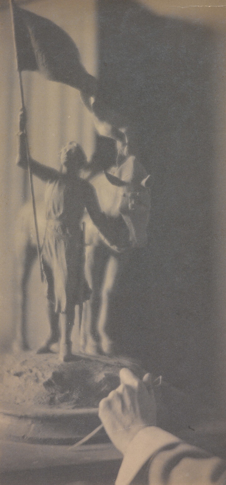A vertically-oriented black-and-white photograph of a hand reaching toward a sculpture of a girl holding a flag in one hand and a horse's rein in the other.