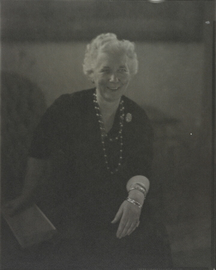 A black-and-white photograph of a smiling White woman with short white hair, seated; one hand holds a book, the other rests on her knee.
