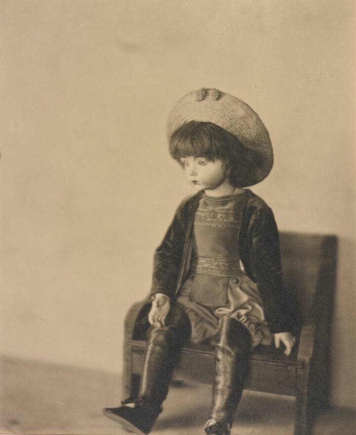 A sepia-toned photograph of a light-colored doll wearing a hat and boots sitting on a small bench.