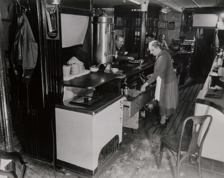 A black-and-white photograph of a woman working in a restaurant while people eat at a counter.