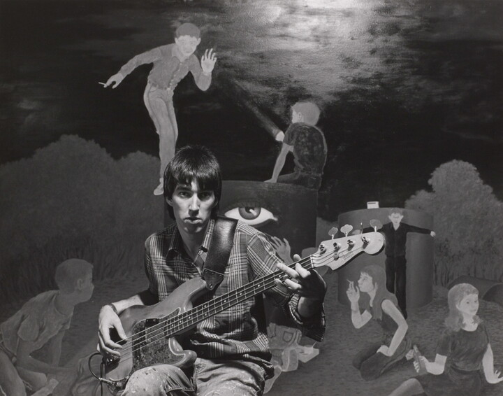 A black-and-white photograph of a young White man playing an electric guitar in front of a mural of people smoking and drinking.