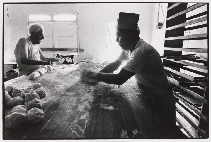 A black-and-white photograph of two men rolling dough on a long table covered in flour.