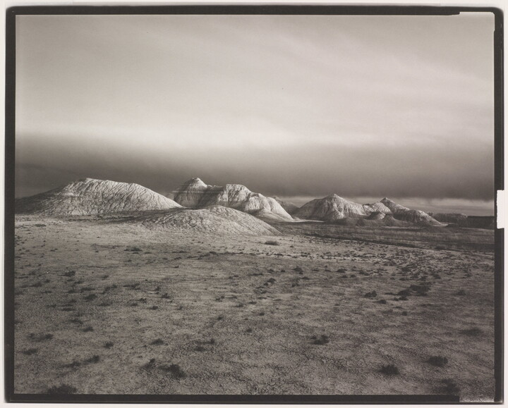 A black-and-white landscape photograph of distant hills at the edge of a flat desert under a dark sky.