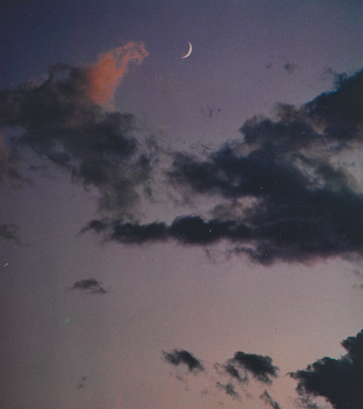 A color photograph of a crescent moon at dusk above thin, dark clouds.