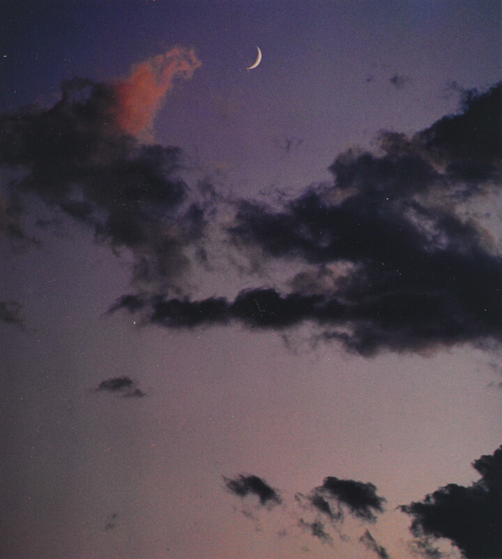 A color photograph of a crescent moon at dusk above thin, dark clouds.