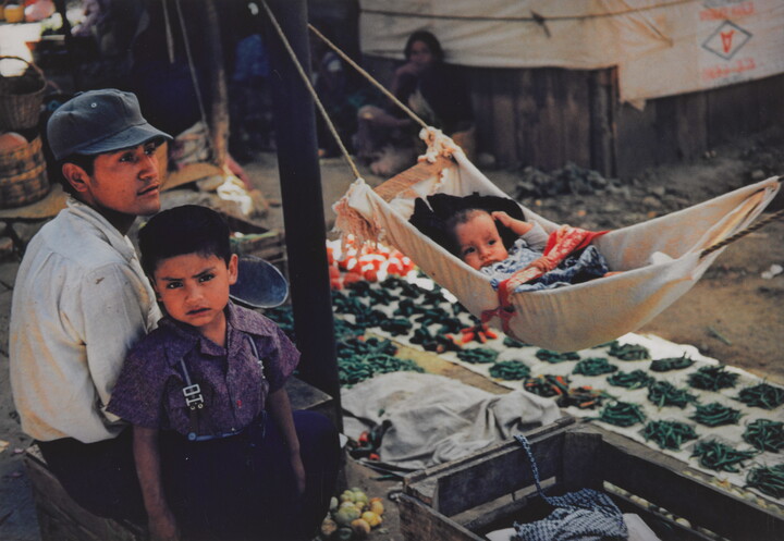 A color photograph of a medium-skinned man, child, and baby; the man seated wearing a cap, child standing next to him as the baby lies in a hammock in front of them.