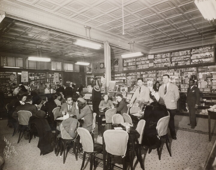 A black-and-white photograph of the interior of a soda fountain filled with a crowd of men and woman socializing.