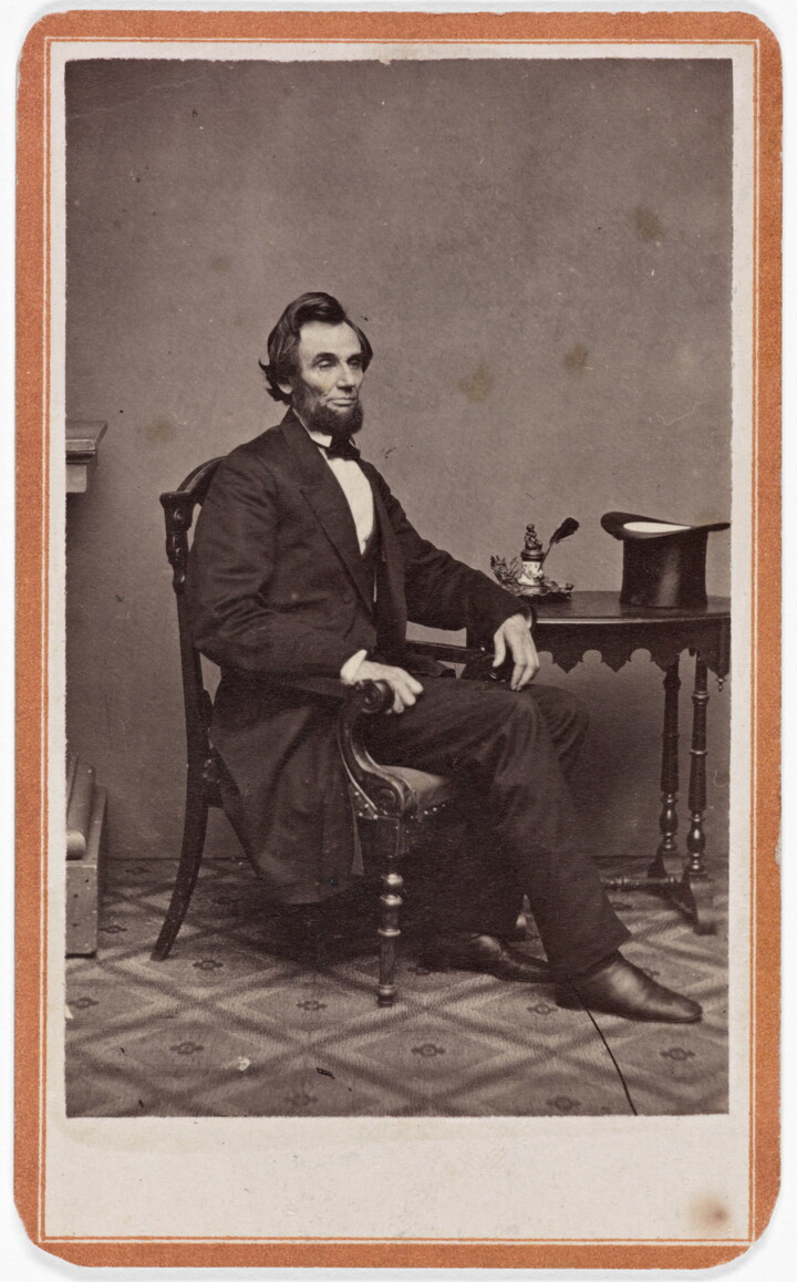 A black-and-white studio portrait photograph of a White man seated, three-quarter view, next to a side table on which sits his stovepipe hat.