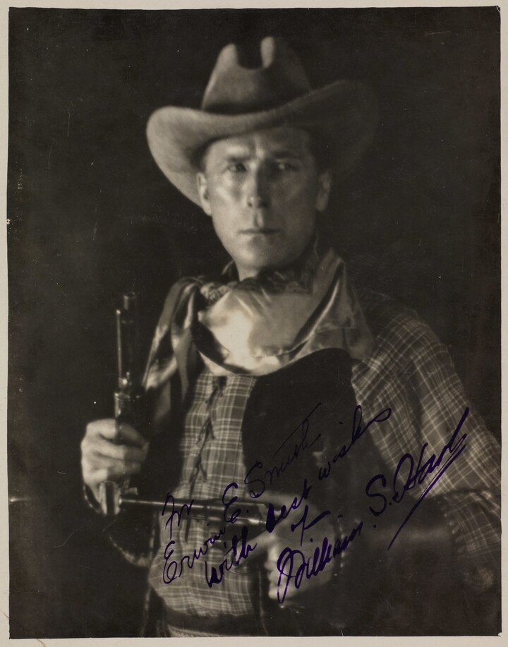 A black-and-white photograph of a White man in western attire holding a pistol in each hand, an autograph in blue in across the bottom.