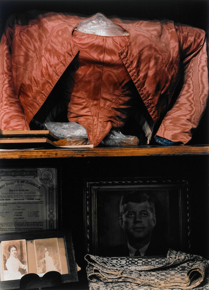 A color photograph of a shelf displaying a red shirt and jacket, photographs of two women, a portrait of John F. Kennedy, some folded fabric, and a Boy Scouts of American certificate.