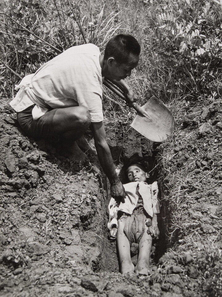 A black-and-white photograph of an Asian man holding a shovel crouched down next to a freshly dug grave containing a emaciated body.